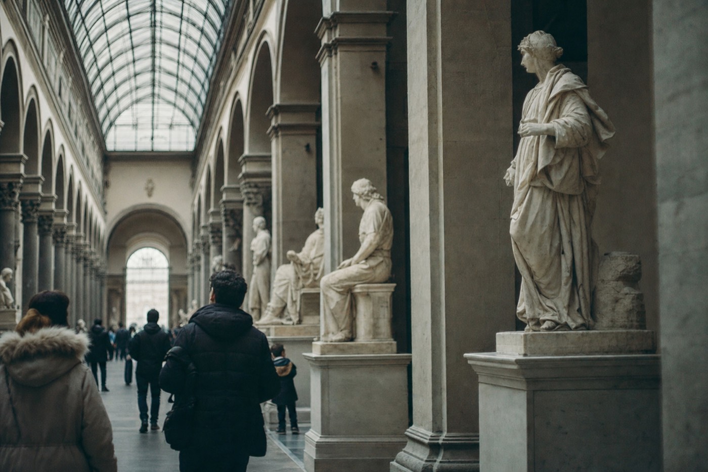 Long sculpture corridor of the Accademia Gallery in Florence with marble figures lit by a glass skylight
