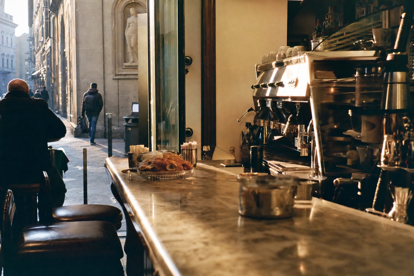 A marble-topped Florentine bar counter at dawn with an espresso machine and pastries near Via Ricasoli close to the Accademia