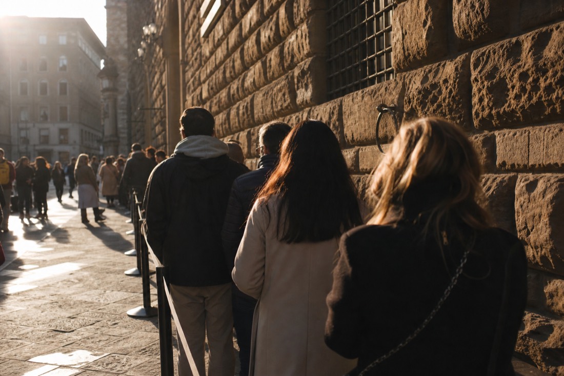 A long line of visitors waiting under morning sun along a Florence street beside a stone gallery wall