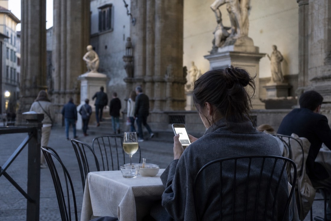 A traveller booking a timed museum slot on a phone at a Florence cafe with the Accademia stonework behind
